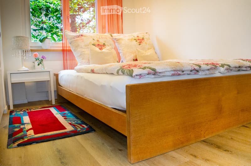 Cozy bedroom with a wooden bed, floral bedding, a nightstand with a lamp, and a colorful rug on the floor.