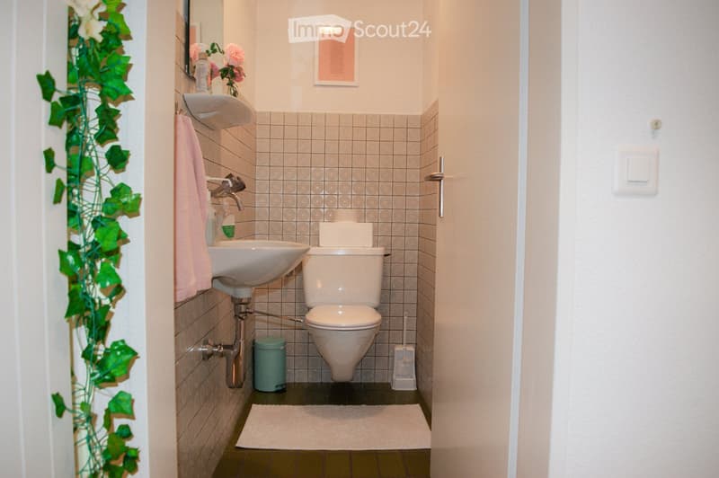 A compact bathroom with a white sink, toilet, and a pink towel hanging on the wall. The walls are tiled with a light gray pattern, and a small white rug is placed on the floor. A flower vase with pink flowers sits on the sink, and a mirror is mounted abov