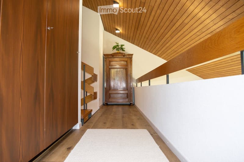 Wooden hallway with a staircase, wooden cabinet, and small plant on top. The ceiling is wooden with recessed lighting, and the walls are white.