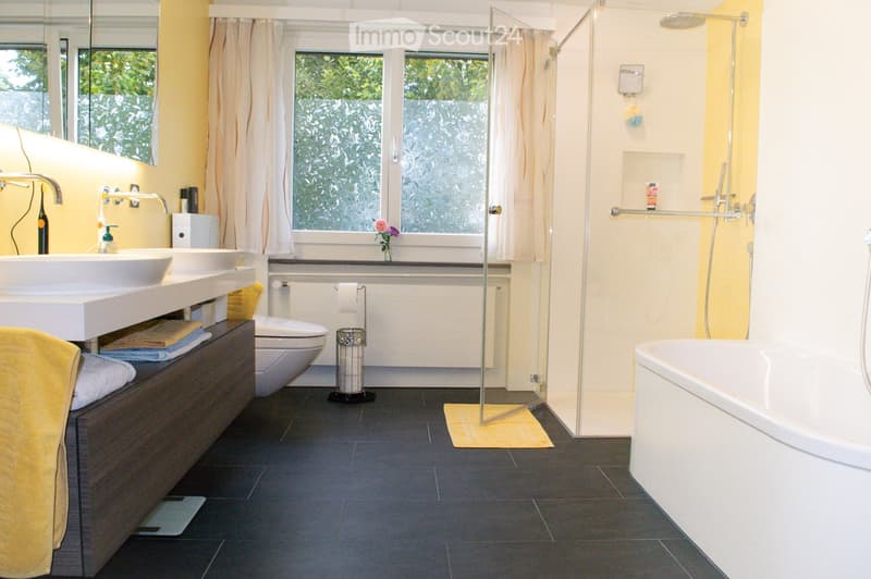 Modern bathroom with double sink, toilet, bathtub, shower, and large window with frosted glass. Features dark tiled floor, light yellow walls, and a wooden cabinet with towels.