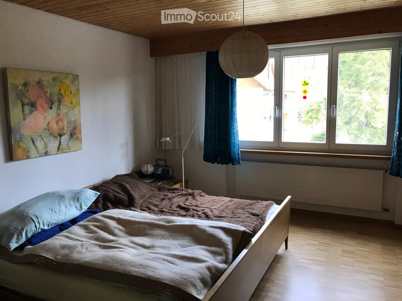 Cozy bedroom with wooden floor, large window with blue curtains, bed with brown and white bedding, nightstand with a lamp, painting on the wall, and a hanging spherical light fixture.