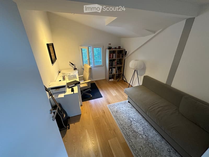 A cozy, multi-functional room with a sofa, desk, bookshelf, and guitar, featuring wooden flooring and large windows.