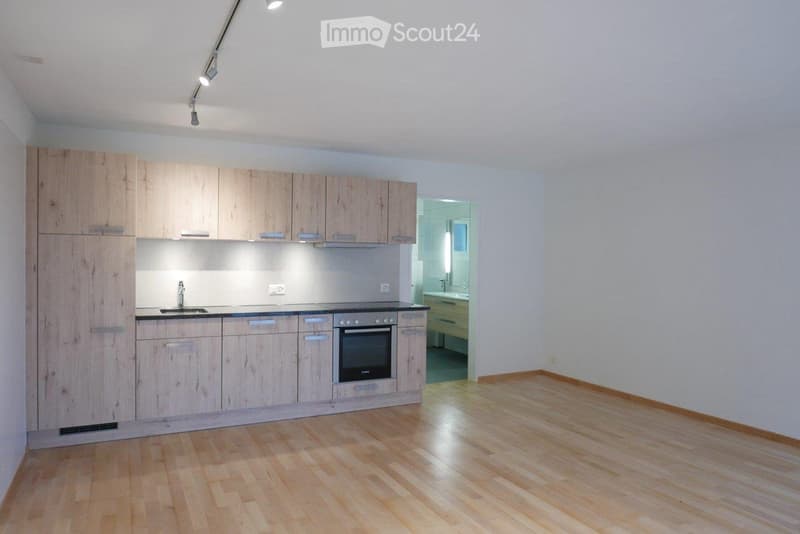 Empty room with wooden flooring, white walls, and a fitted kitchen area with cabinets and appliances visible in the background.