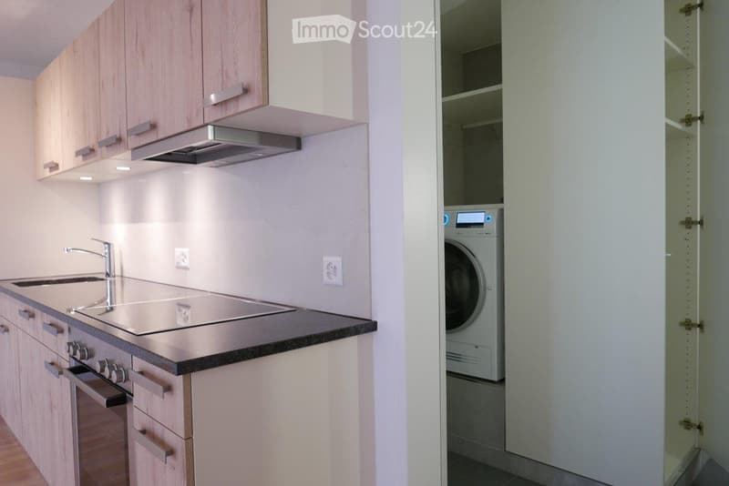 Modern kitchen with light wooden cabinets, stainless steel sink, faucet, oven, and range hood. Adjacent laundry area with a washing machine.