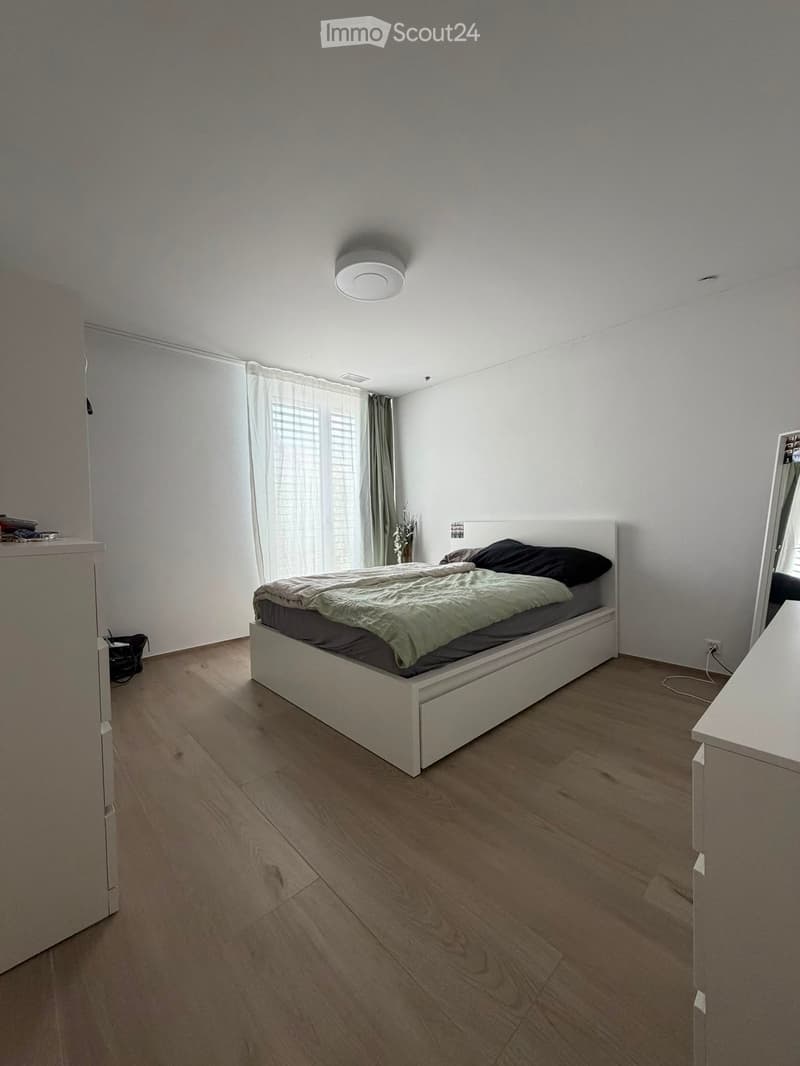 Modern bedroom with wooden flooring, white walls, large window with blinds, bed with dark bedding, and a mirror on the right side.