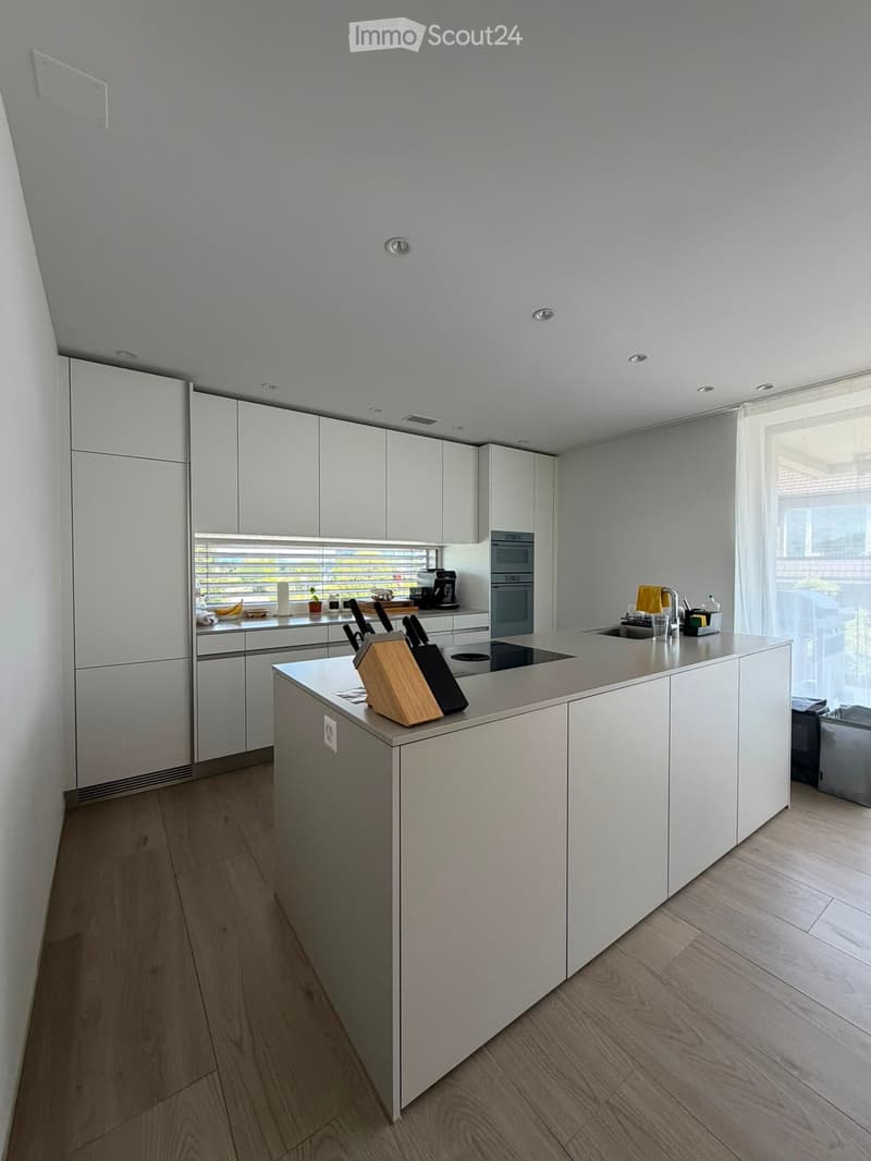 Modern kitchen with white cabinets, a central island, and stainless steel appliances. The island features a knife block and a sink with a modern faucet. The countertops are light-colored, and the floor is made of light wood.