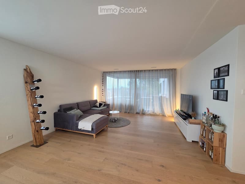 Modern living room with light wooden flooring, grey sectional sofa, ottoman, coffee table, TV, wine rack, and picture frames on the wall.
