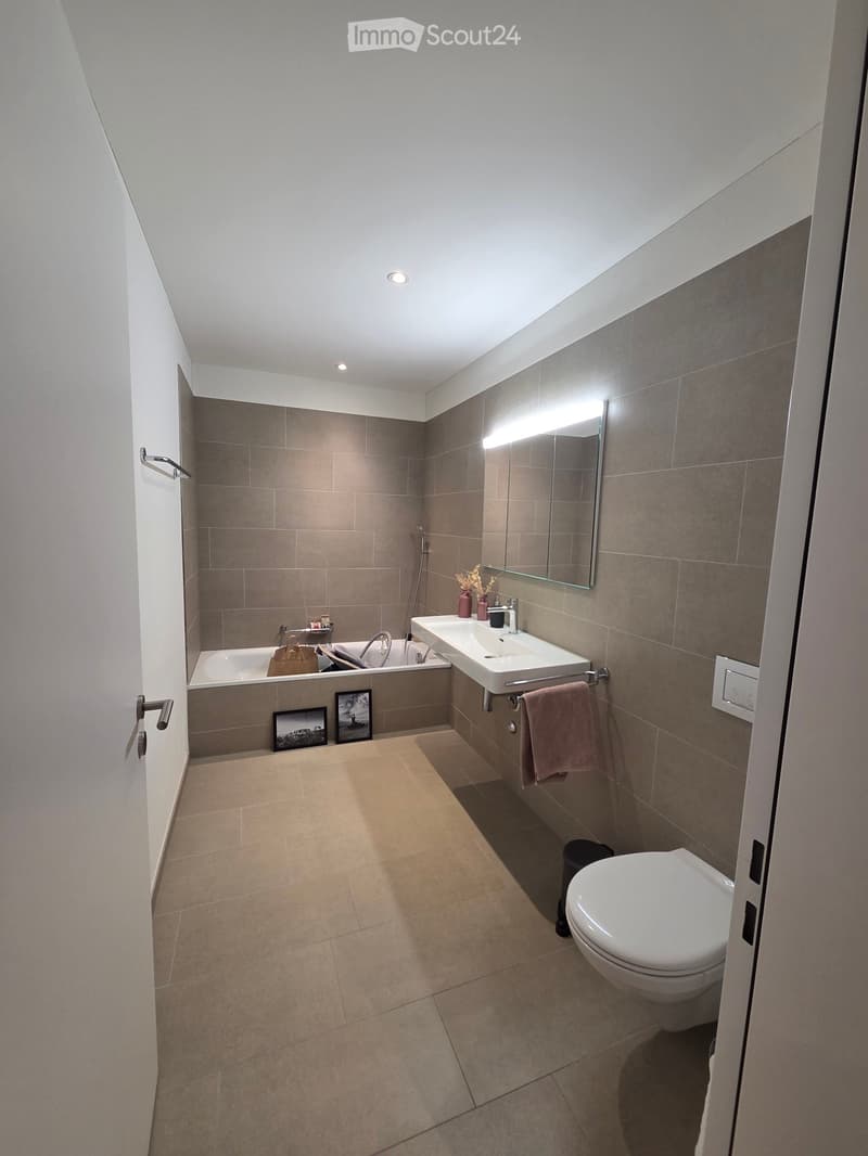 Modern bathroom with a bathtub, sink, toilet, mirror, and two picture frames on the floor. Beige tiles and grey walls.