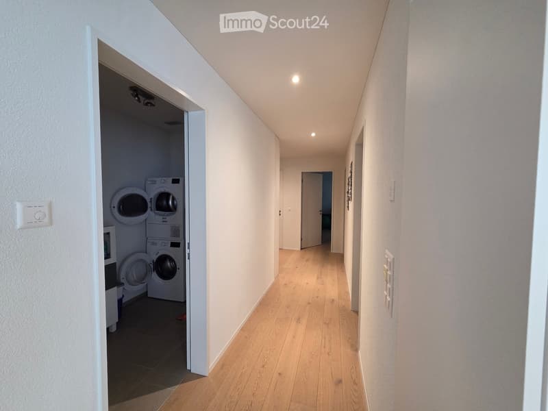 A narrow hallway with light wooden flooring and white walls, featuring a small laundry area with stacked washing machines and dryers visible to the left.