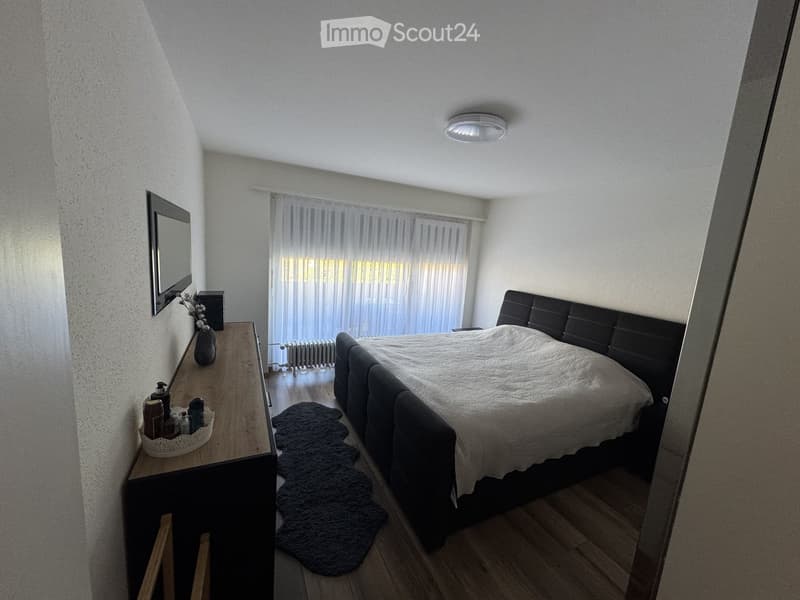 Cozy bedroom with a large bed, nightstand, mirror, vase, and rug. The room features wooden flooring and large windows with sheer curtains.
