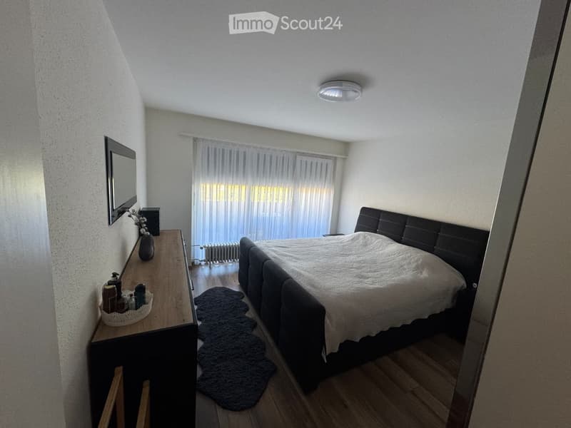 Modern bedroom with a large bed, nightstand, mirror, and a small table with decorative items. The room features wooden flooring and large windows with sheer curtains.