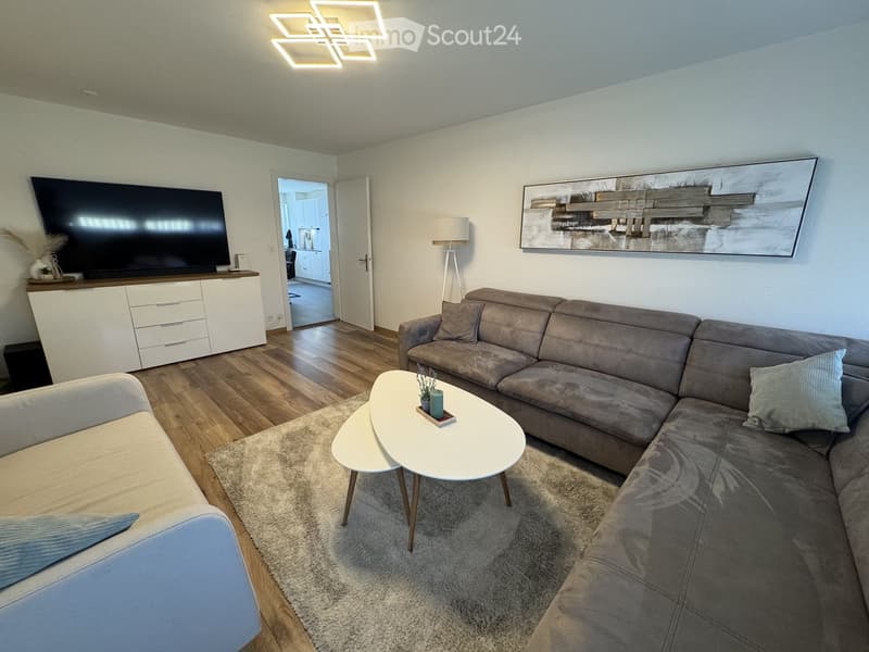 Modern living room with a grey sectional sofa, white coffee table, armchair, TV stand, floor lamp, and a cabinet. The room features wooden flooring and a large abstract painting on the wall.
