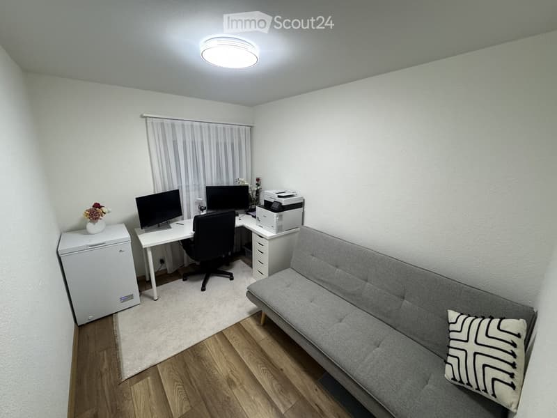 A small, well-lit room with a sofa, desk, chair, printer, and refrigerator. The room features wooden flooring and white walls.