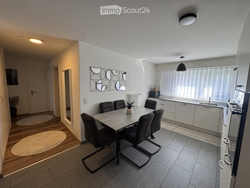 Modern open-plan living and dining area with a dining table, six chairs, a kitchen area with cabinets, sink, microwave, and a decorative mirror on the wall.