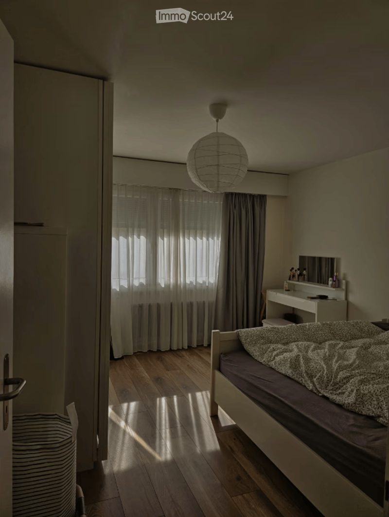 Bedroom. A modern, minimalist bedroom with a wooden floor, a single bed with a patterned duvet, a vanity with a mirror, and a hanging spherical lamp. The room features large windows with sheer curtains allowing natural light to filter in.