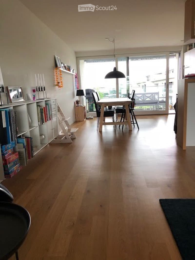 Bright dining area with wooden flooring, large glass doors leading to a balcony, a dining table with chairs, a bookshelf, and decorative items.