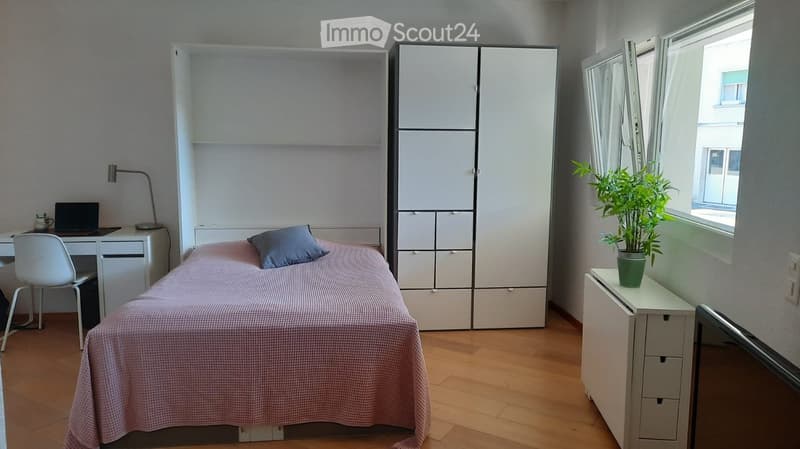 A modern, minimalist bedroom with a single bed, a desk with a laptop, a white wardrobe, and a potted plant near the window.