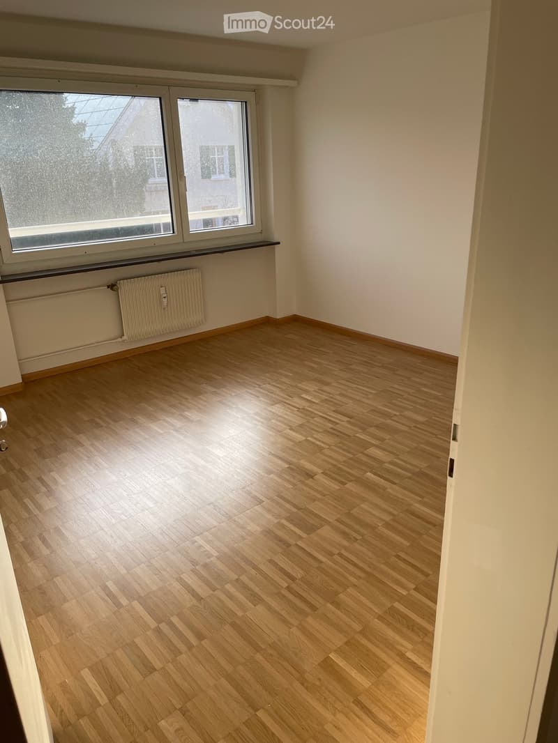 Empty room with light wooden flooring, white walls, large windows, and a radiator under the windows.