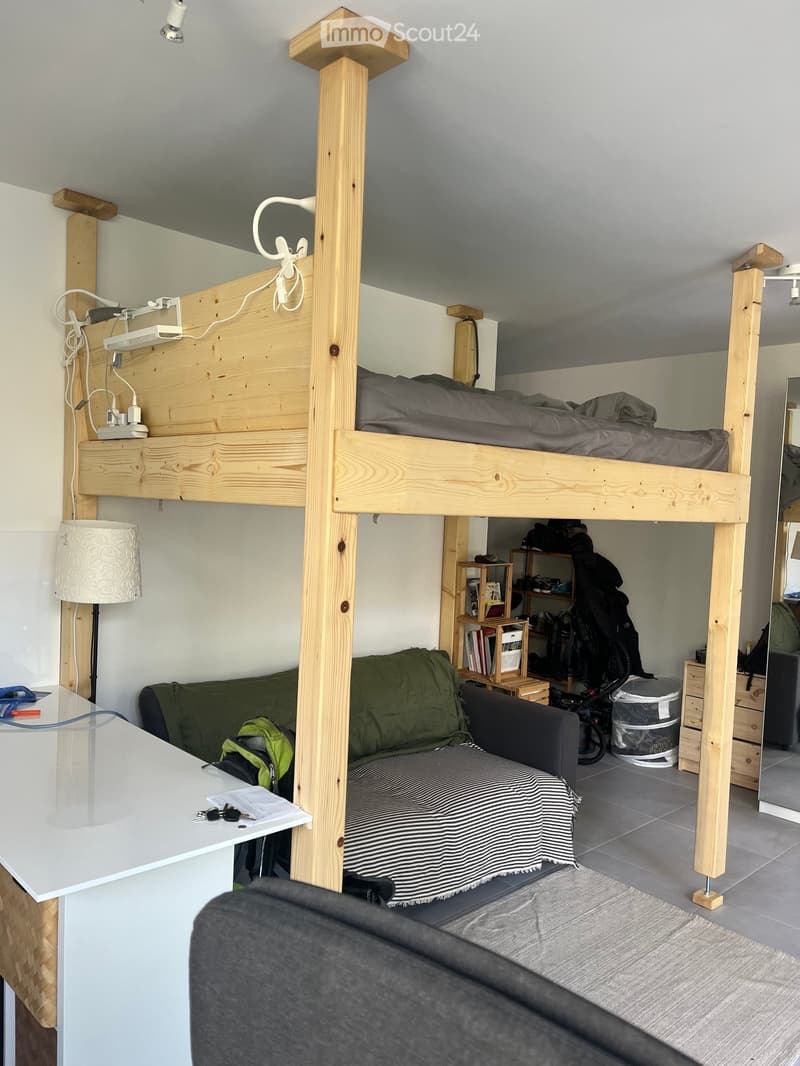 A compact, furnished room with a lofted bed, a sofa, a small table, and shelves. The space includes a lamp, a mirror, and a backpack on the floor.