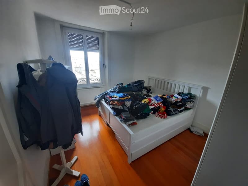 Bedroom with wooden floor, white bed frame, and clothes on the bed. Wardrobe with jackets visible on the left side.