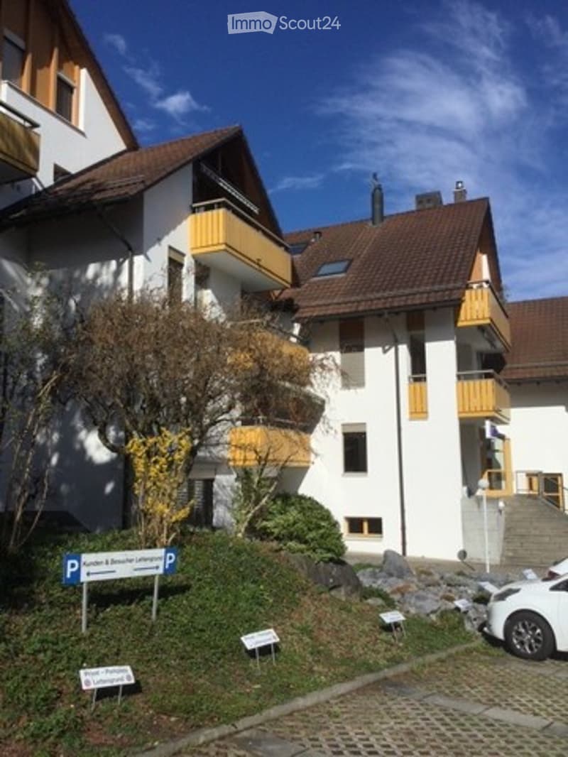 Multi-story residential building with white facade, brown roof, and yellow balconies. A parking sign is visible in the foreground.