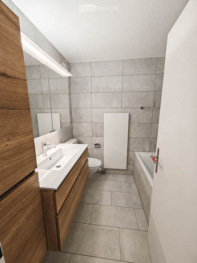 Modern bathroom with a double sink, toilet, bathtub, and a radiator. The walls are tiled in grey, and the floor is tiled in light grey. There is a wooden vanity cabinet next to the sink.