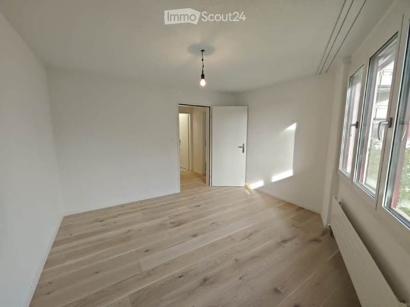 Empty room with light wooden flooring, white walls, and large windows allowing natural light. A single door leads to another room.