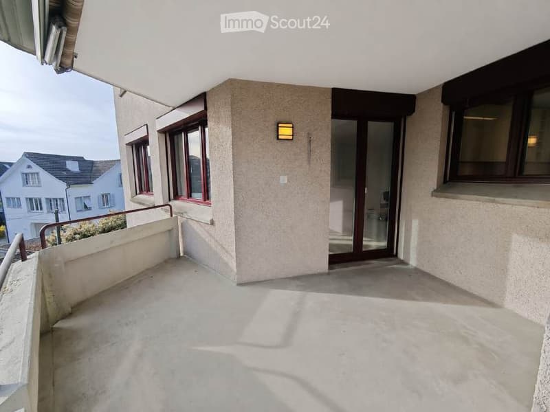 Balcony with concrete floor, metal railing, and glass door leading to interior. View of neighboring houses and greenery.