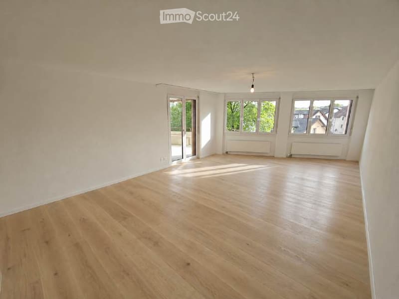 Empty room with light wooden flooring, white walls, large windows, and a sliding glass door leading to a balcony.