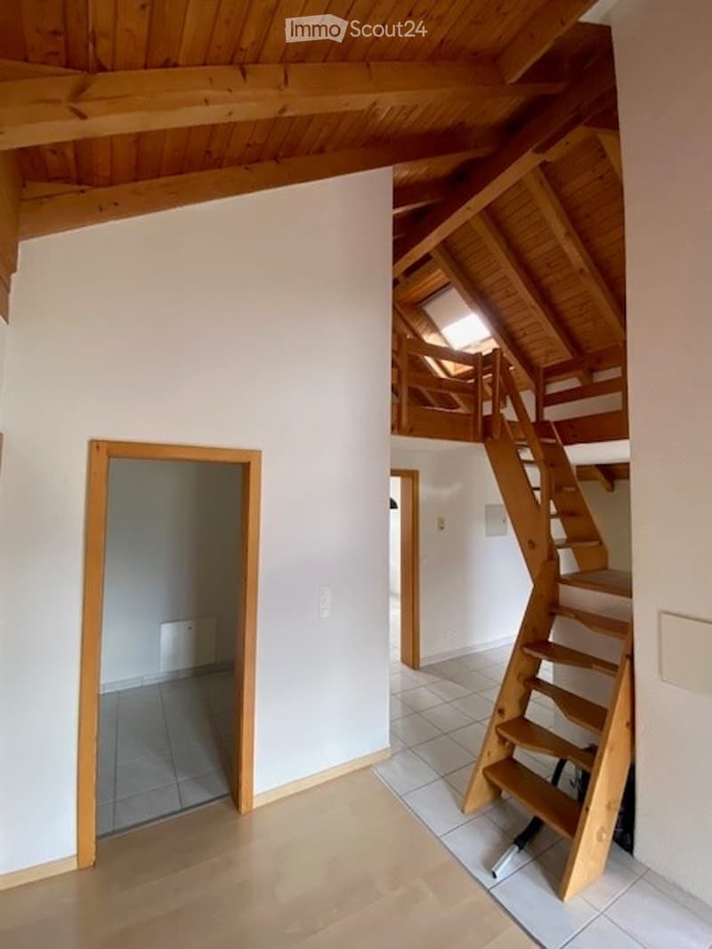 Bright, open-plan space with wooden floors, white walls, and exposed wooden beams. Features a staircase leading to an upper level with a small landing and a window.