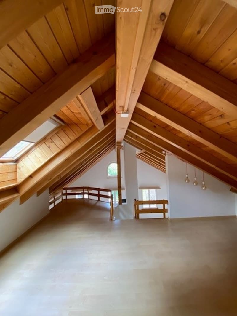 Spacious attic room with exposed wooden beams, skylights, and a small bench. The room is completely empty with white walls and a smooth concrete floor.