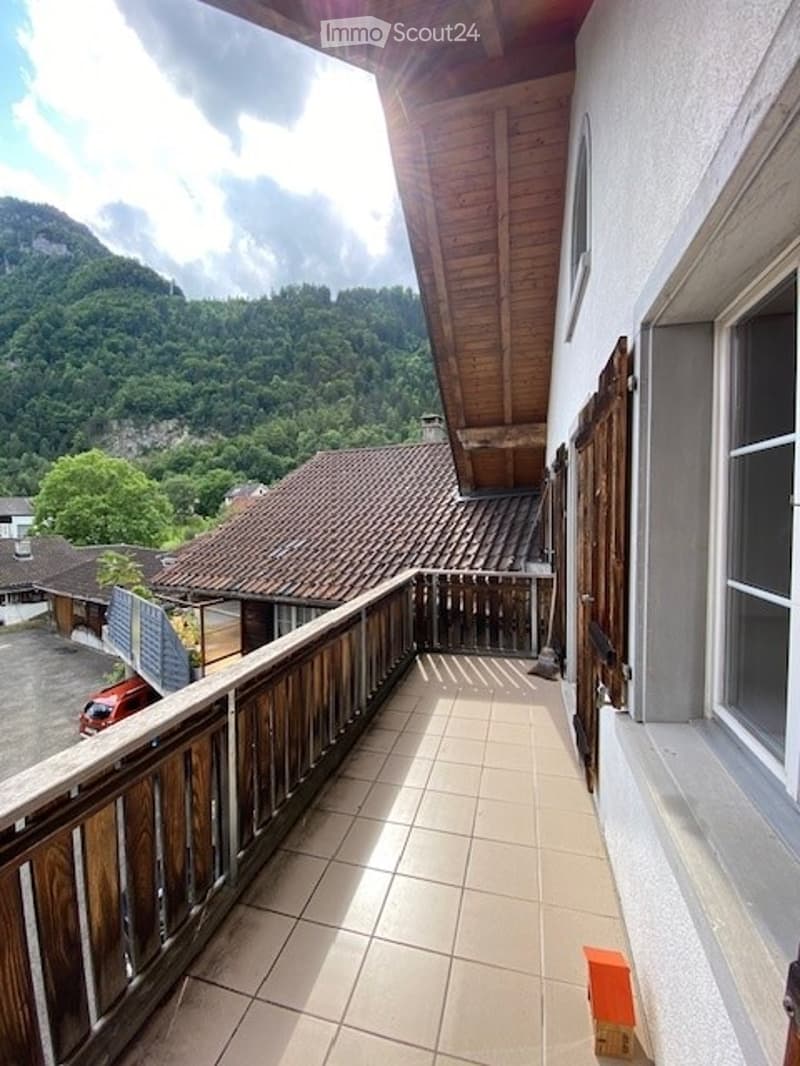 Balcony with wooden railing, tiled floor, overlooking a scenic mountain landscape with houses and trees.