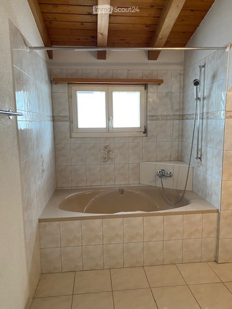 Bathroom with a bathtub, shower, and window. The walls are tiled, and the ceiling has wooden beams.