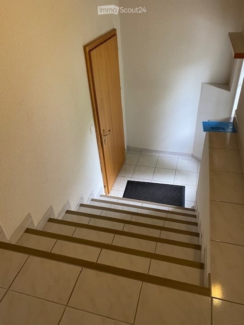 Staircase leading up with wooden handrail, tiled steps, and a small black doormat at the base of the stairs.