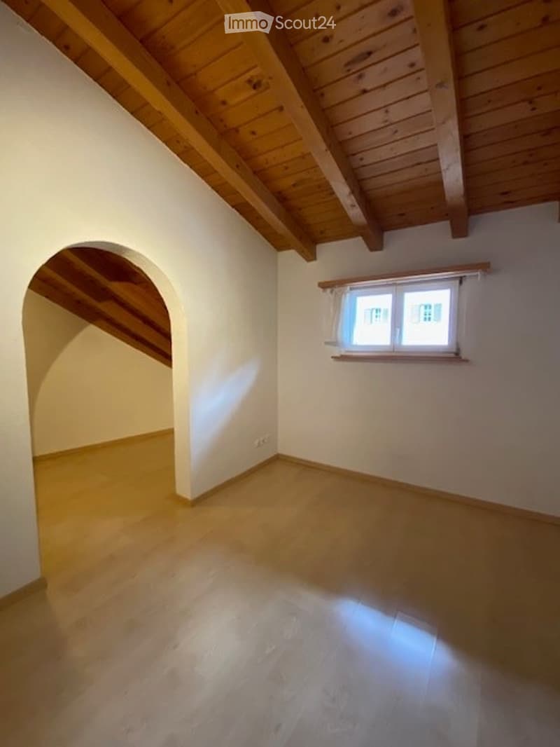 Empty attic room with wooden slanted ceiling, wooden floor, and a single window with white frame and curtains.