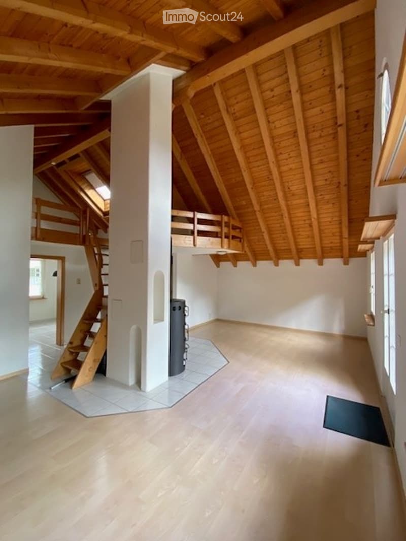 Bright, open attic space with wooden beams and exposed ceiling. Features wooden flooring, a central column, a staircase leading to an upper level, and large windows allowing natural light.