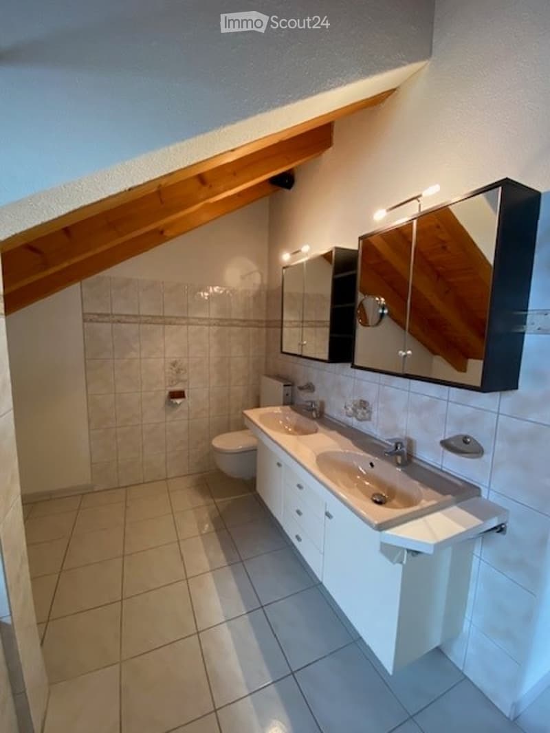 Modern bathroom with a double sink, large mirror, toilet, and white cabinets under the sinks. The room features a sloped wooden ceiling and tiled walls and floor.