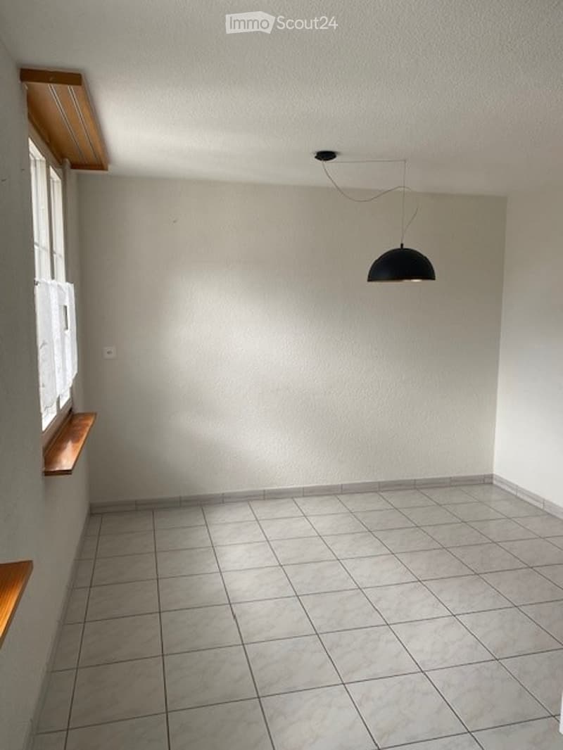 Empty room with tiled floor, white walls, and a single pendant light fixture. There are wooden window sills and a small wooden shelf on the left side.