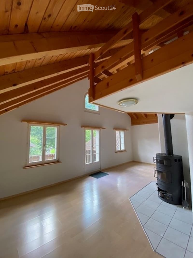 Bright, empty room with wooden ceiling beams, large windows, and a tiled area near a wood-burning stove. The floor is light wood, and the walls are white.