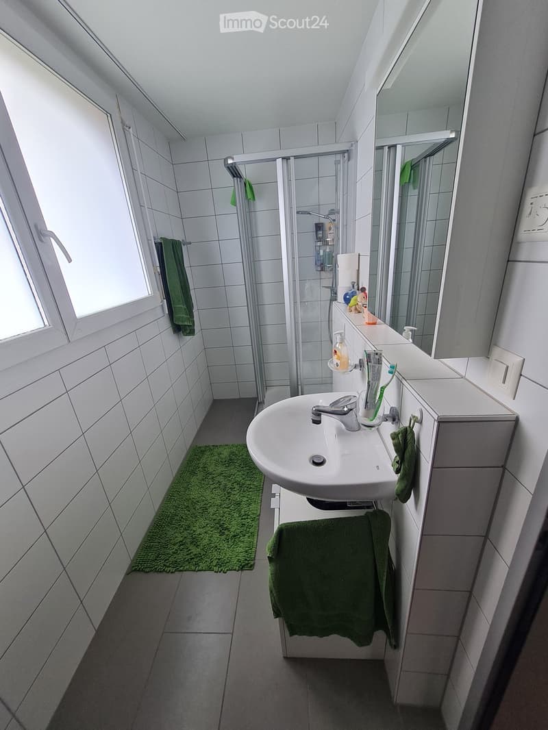 Modern bathroom with white tiles, a sink with a chrome faucet, a large mirror, a shower area with a glass door, and green towels on the cabinet and floor.