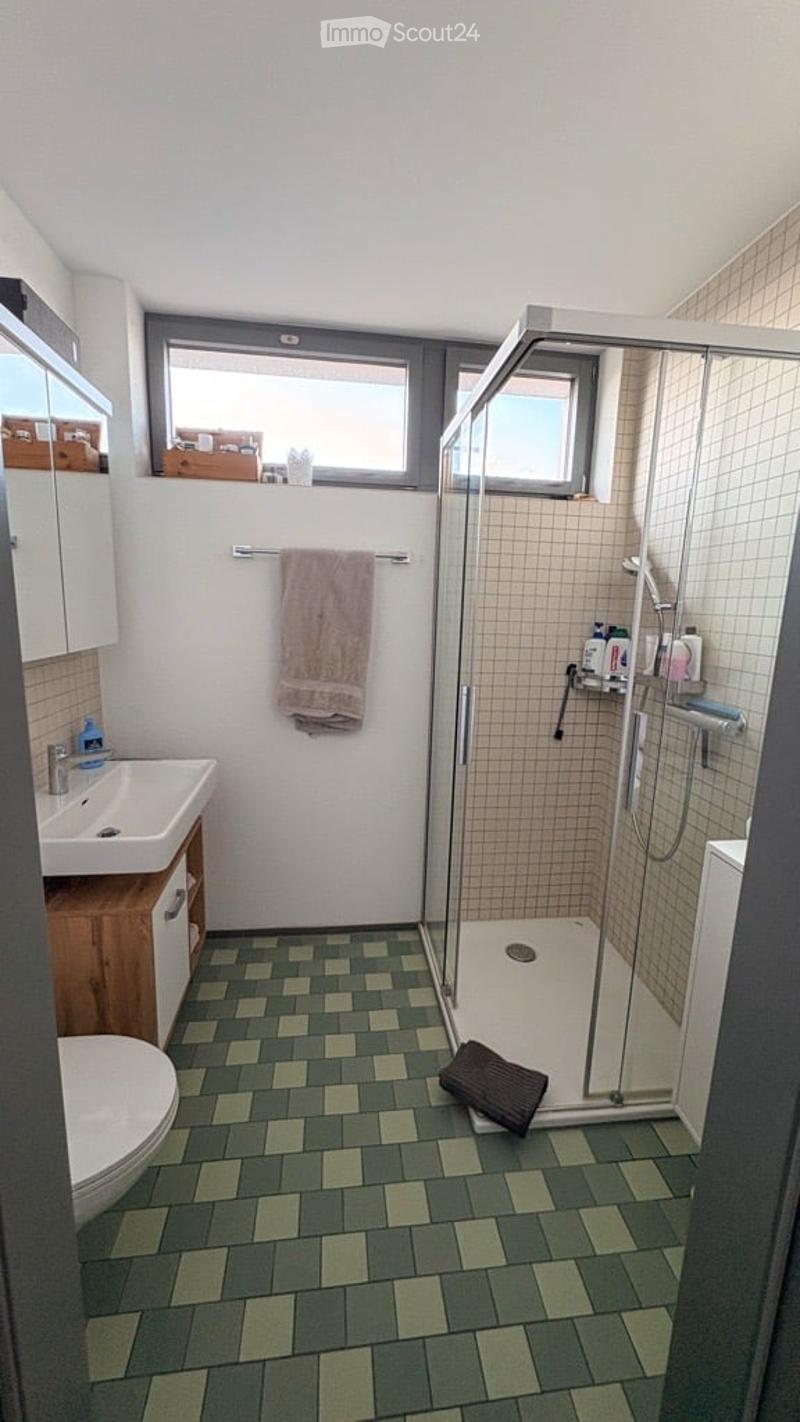 Modern bathroom with green and white checkered tiles, a glass-enclosed shower, a white sink with a wooden cabinet, and a toilet. A towel hangs on a rack, and a mirror is mounted on the wall.