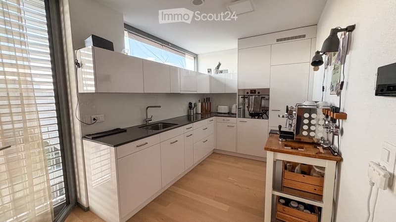 Modern kitchen with white cabinets, black countertops, wooden island, coffee machine, and large window with blinds.