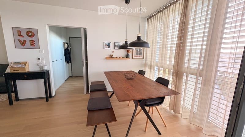 Bright dining area with wooden floor, large window with curtains, modern dining table with chairs, bench seating, and decorative wall art.