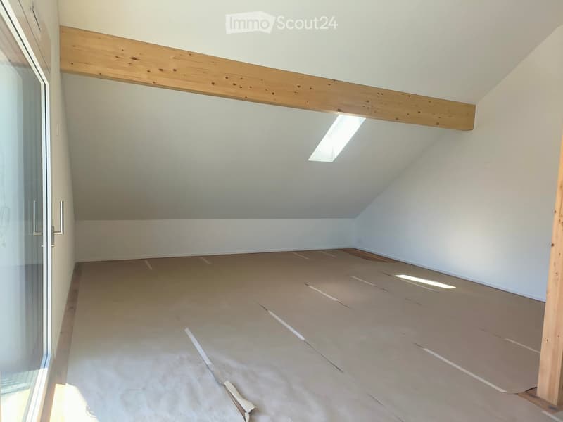 Empty attic room with exposed wooden beams, white walls, and a skylight window. The floor is covered with protective cardboard sheets.