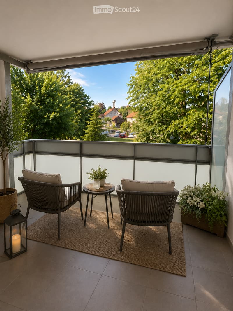 Balcony with two wicker chairs, a small round table, a lantern with a candle, and potted plants, offering a view of greenery and houses.