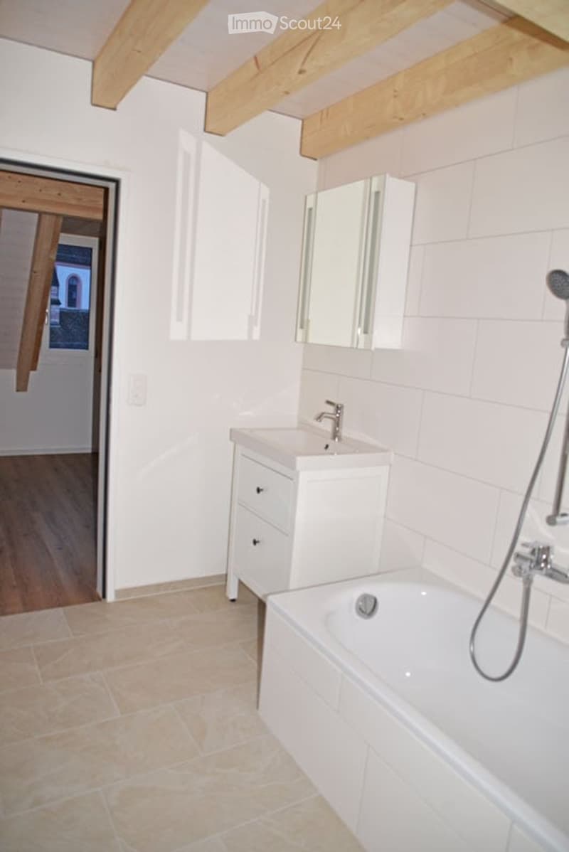 Modern bathroom with white tiles, a bathtub, a sink with a cabinet, and a mirror above it. The room features exposed wooden beams on the ceiling and a wooden floor in the adjacent area.
