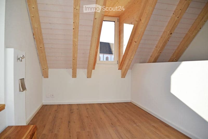 Empty attic room with wooden beams, wooden floor, white walls, and small windows.