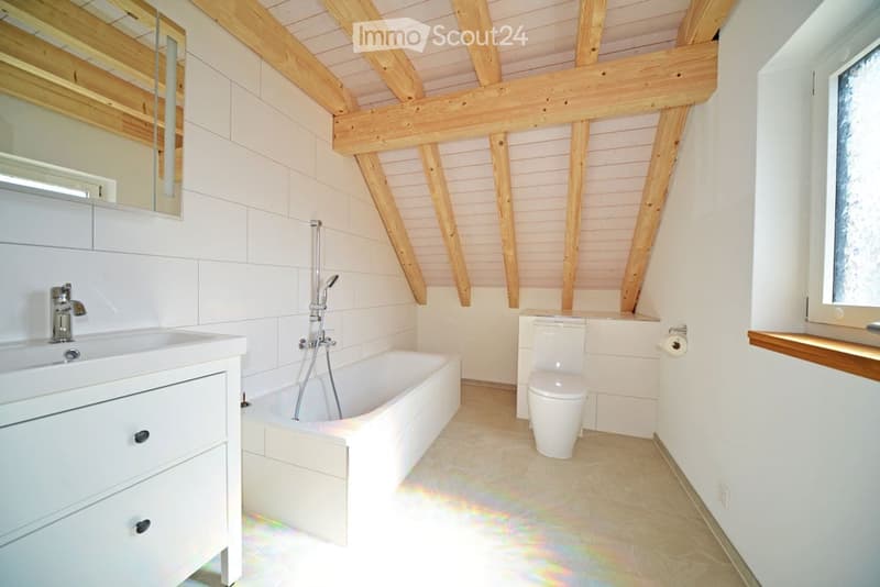 Bright bathroom with white tiles, wooden beams, a bathtub, sink, and toilet. Natural light from a window illuminates the space.
