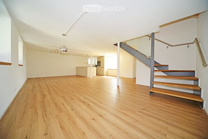 Open-plan unfurnished space with wooden flooring, white walls, and a staircase leading to an upper level. The area includes a small kitchen area in the background.