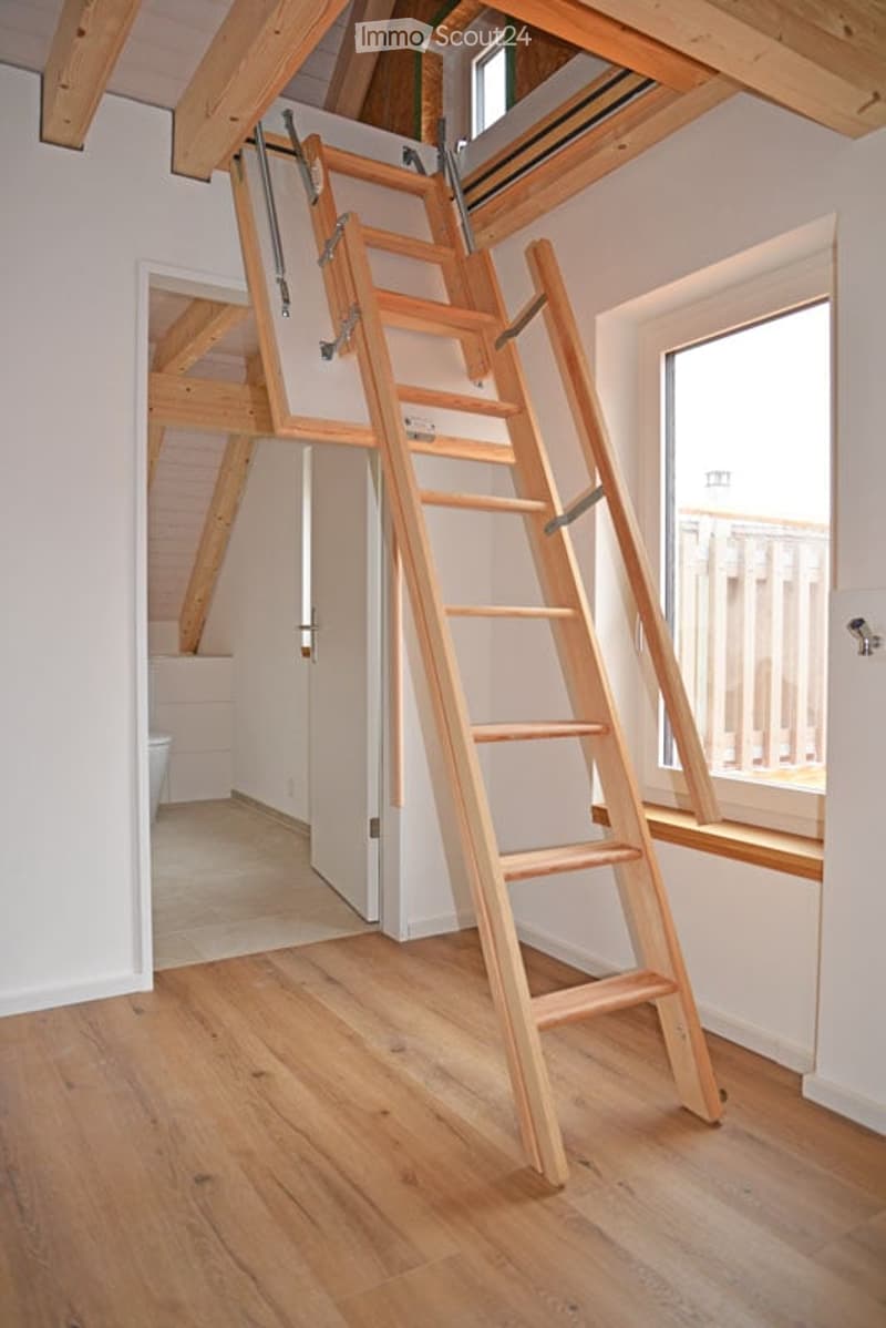 An empty room with wooden flooring and exposed wooden beams on the ceiling. A wooden ladder is positioned against the wall, leading to an attic space. The room has white walls and large windows allowing natural light to enter.